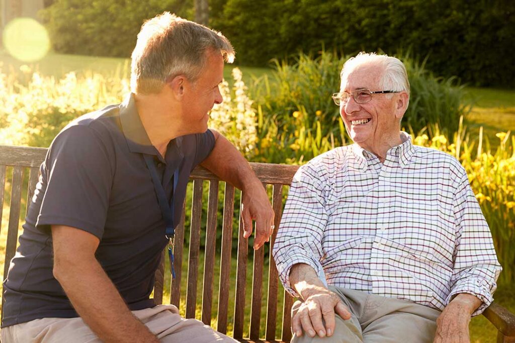 A senior resident in an armchair using a tablet with a caregiver offering friendly guidance nearby.